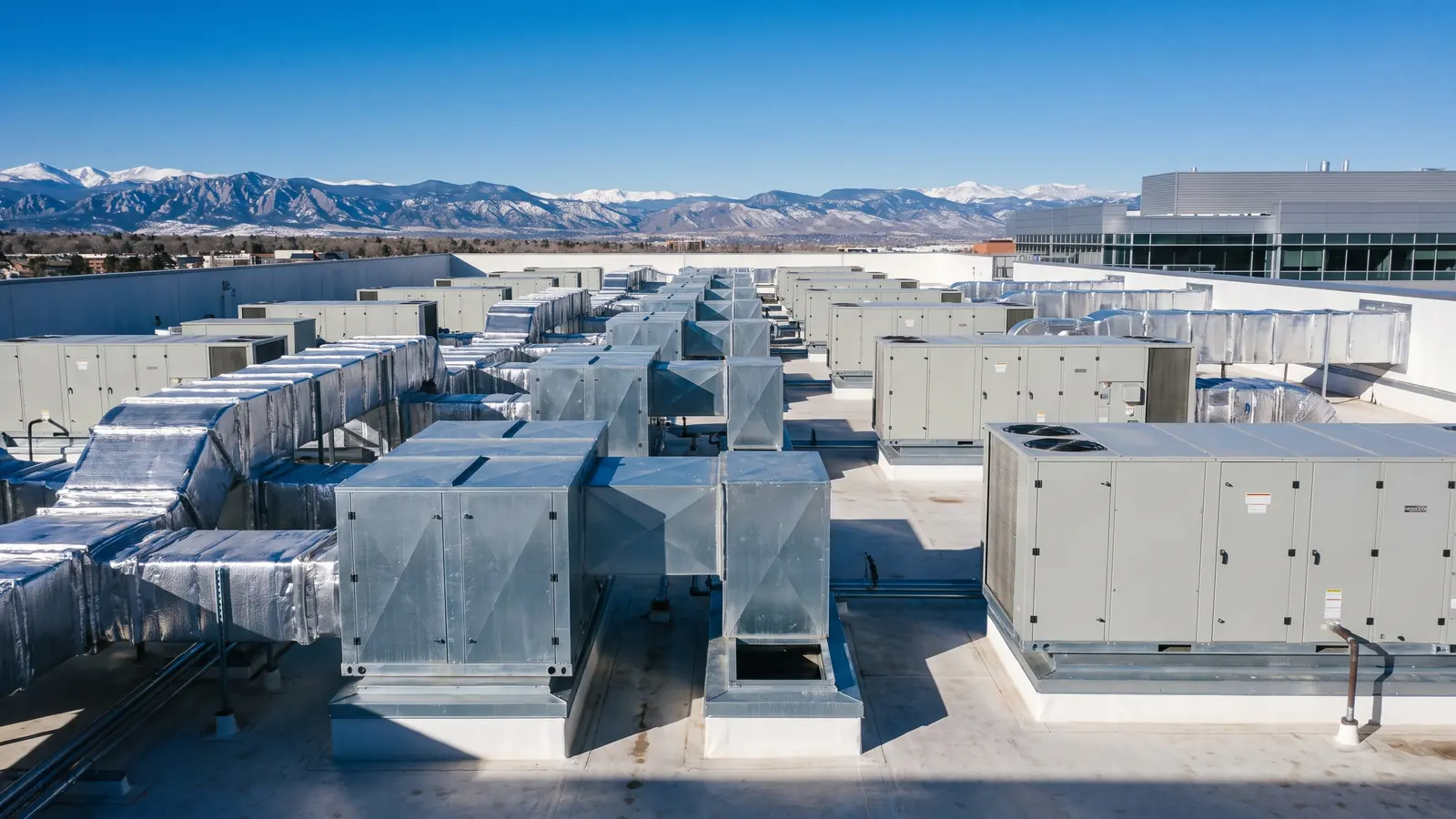 Rows of packaged rooftop HVAC units and insulated ductwork on a commercial roof in Denver, with the snow covered Front Range in the distance